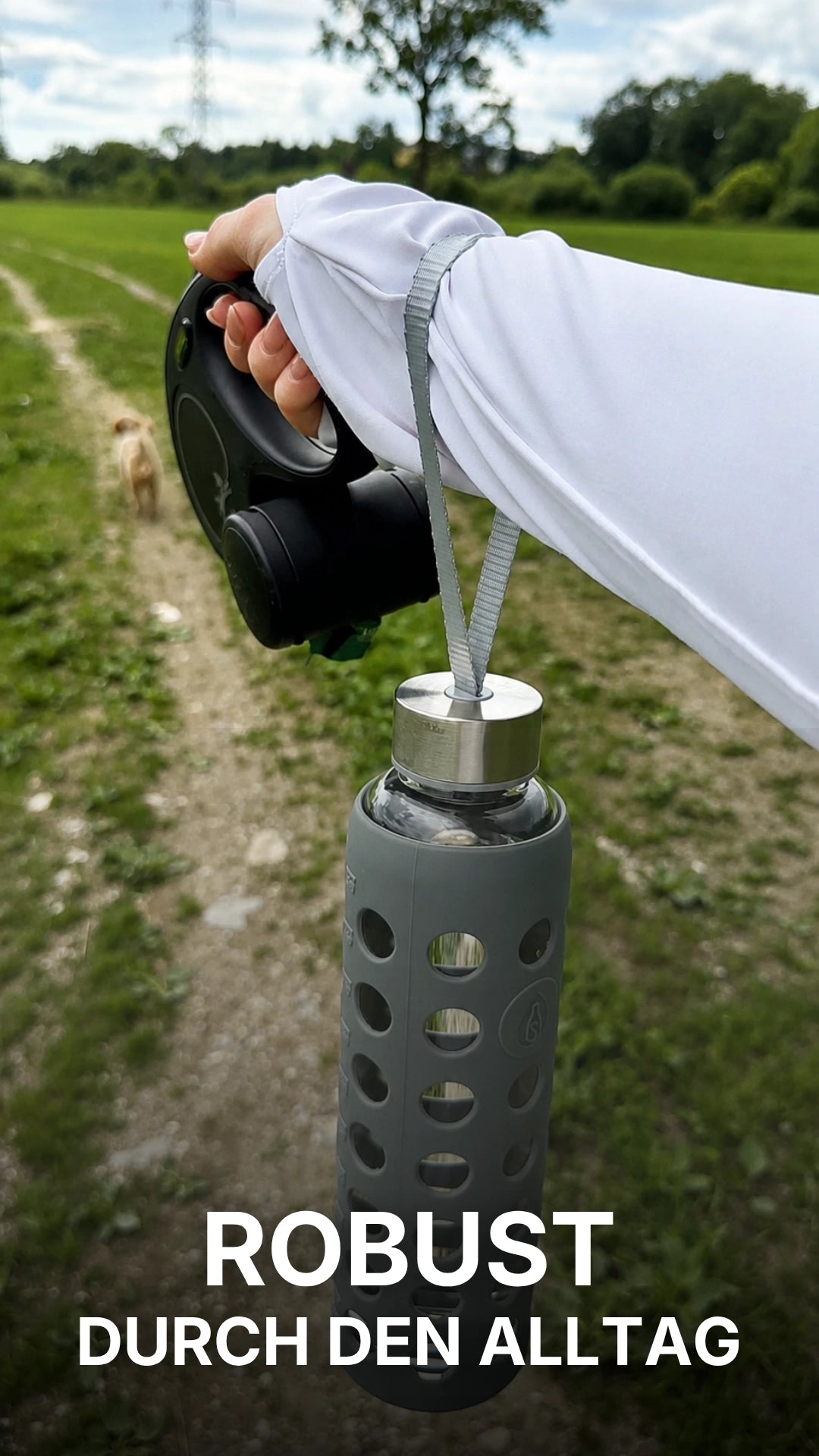 Video laden: Eine junge Frau zeigt auf einem Feldweg eine spottle Glasflasche mit grauer Silikonhülle und Trageschlaufe und berichtet, warum sie die Flasche als robust und sicher im Alltag empfindet. 